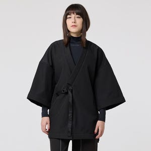 May include: A black kimono jacket with a wrap closure and a matching belt. The outfit includes black trousers with white stitching and black boots. The model is wearing a black polo neck. The overall aesthetic is minimalist and modern.