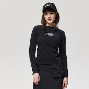 May include: Black long-sleeve shirt with a logo, paired with a black skirt-pants hybrid and combat boots. The outfit includes a black baseball cap. The ensemble is a modern, urban style.