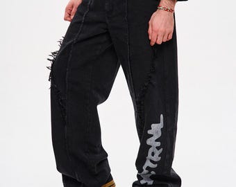 May include: Black denim jeans with distressed detailing and a wide leg fit. The jeans feature a silver graphic on the lower leg and are paired with black sneakers with brown soles. The model is wearing a black long-sleeve shirt.