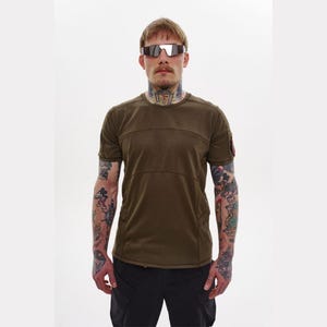 Può includere: T-shirt verde oliva a maniche corte con dettagli a pannelli orizzontali sul petto. La maglietta ha una piccola toppa sulla manica sinistra ed è abbinata a pantaloni cargo neri. La persona nell'immagine indossa occhiali da sole.
