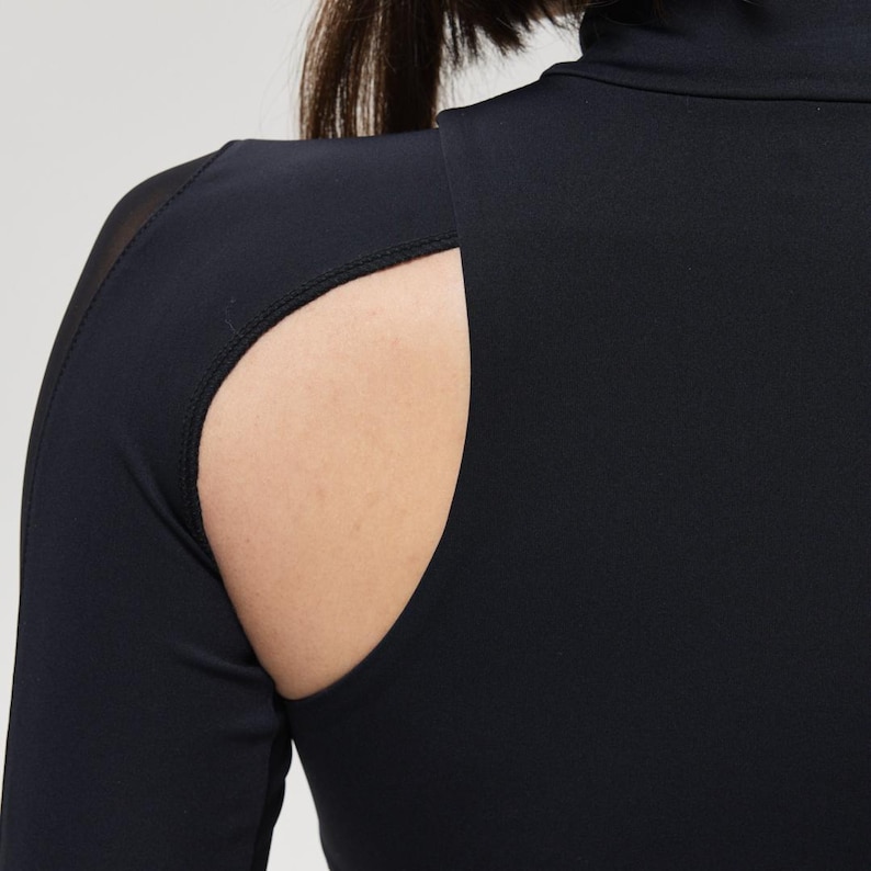 May include: Black long-sleeve top with a high neckline and a cutout detail on the shoulder. The top is made of a smooth, stretchy fabric. The cutout reveals a portion of the shoulder and upper arm.