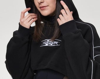 May include: Black cropped hoodie with white piping and the text "TUR WEAR" in white. The hoodie has a drawstring hood and long sleeves. The model is wearing a black turtleneck and a black belt.