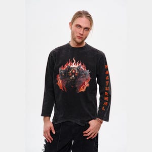 Könnte beinhalten: Schwarzes Langarmshirt mit Used-Look. Das Shirt zeigt eine Grafik einer Katze, die auf der Vorderseite von Flammen umgeben ist. Das Wort "NATURAL" ist vertikal in Rot auf dem linken Ärmel aufgedruckt.