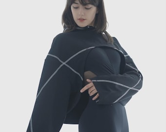 May include: A black top with white stitching and a high collar, worn with black leggings. The stitching forms a geometric design on the shoulders and sleeves. The outfit is a modern, sleek design. The model is wearing a black outfit.