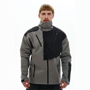 Könnte beinhalten: Grau-schwarze Jacke mit hohem Kragen und mehreren Reißverschlusstaschen. Die Jacke hat ein einzigartiges Design mit einem kontrastierenden schwarzen Einsatz auf der rechten Seite. Die Jacke ist durchgehend mit Reißverschluss versehen und hat schwarze Akzente an den Ärmeln.