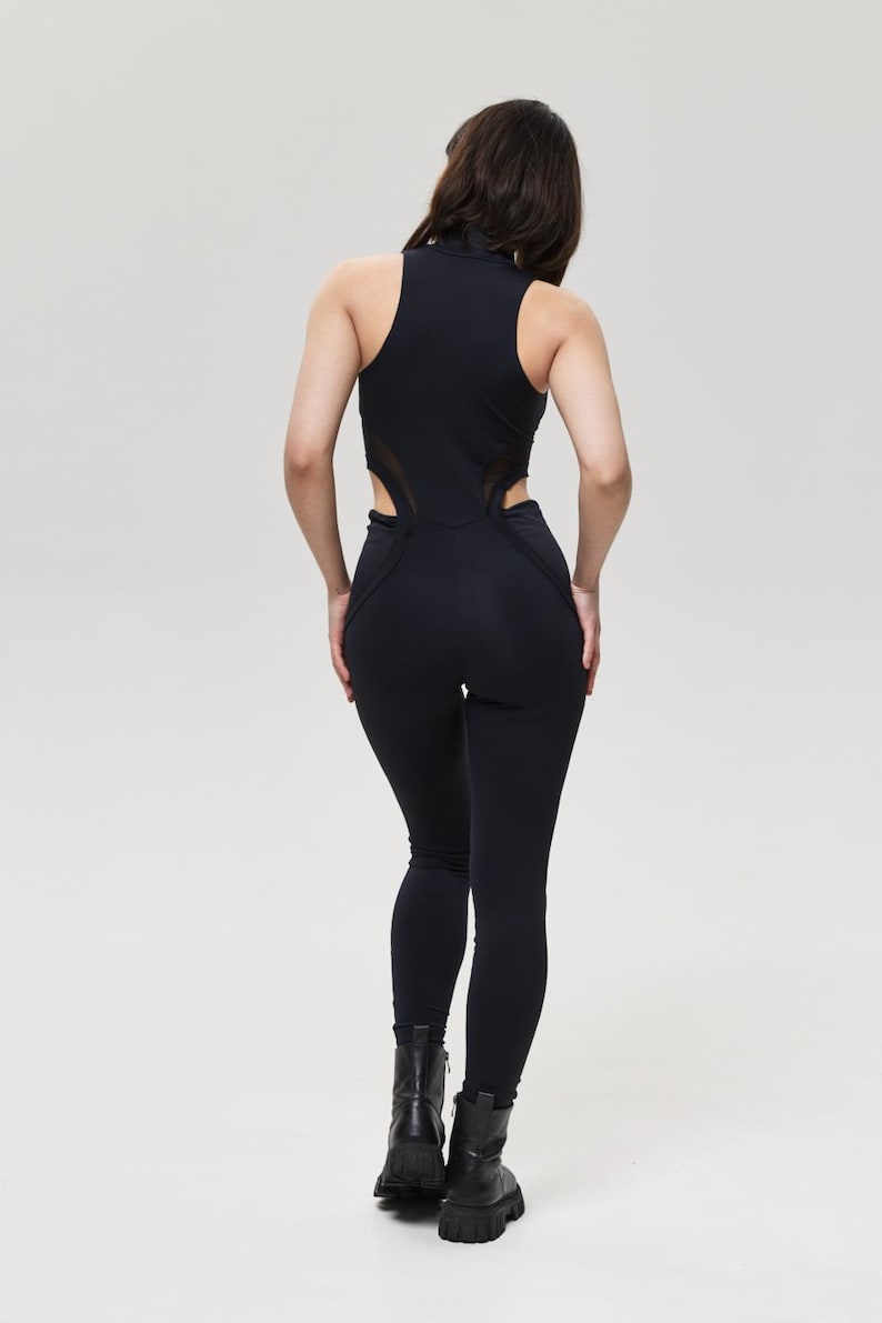 May include: Black jumpsuit with a high neckline and cut-out detailing on the sides. The jumpsuit features a sleeveless design and fitted leggings. The model is wearing black boots. The background is a solid light gray.