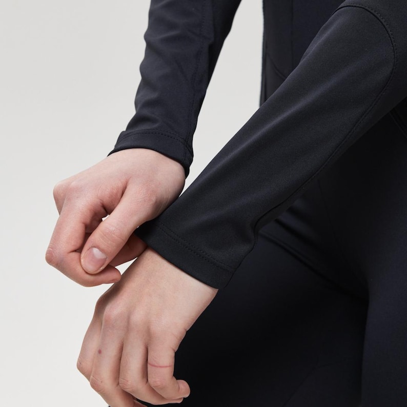 May include: Black long-sleeved athletic top. The sleeves are fitted and end at the wrist. The fabric appears to be a smooth, stretchy material. The top is paired with black athletic pants. The background is a solid, neutral color.