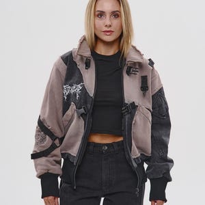 Op de afbeelding: Een patchwork jas in grijstinten en taupe met zwarte accenten. De jas heeft meerdere zakken, gespen en een ritssluiting. Het ontwerp bevat geborduurde tekst en een distressed look, wat een unieke, edgy uitstraling creëert.