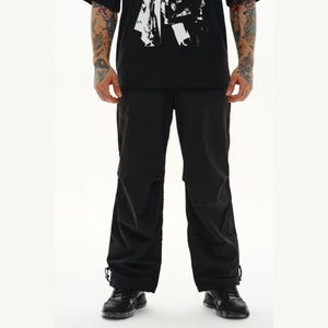 May include: Black pants with a relaxed fit, featuring adjustable drawstrings at the ankles. The pants are made of a matte material and have a simple, streamlined design. The pants are worn with black shoes and a black t-shirt.
