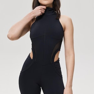 May include: Black jumpsuit with a high neckline and halter-style top. The jumpsuit features mesh cutouts on the sides and a form-fitting design. The model is wearing the jumpsuit. The jumpsuit is made of a stretchy material.