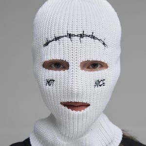 May include: White knitted balaclava featuring black barbed wire embroidery above the eyes. The words "NOT" and "NICE" are embroidered on either side of the mouth opening. The balaclava covers the entire head and neck.