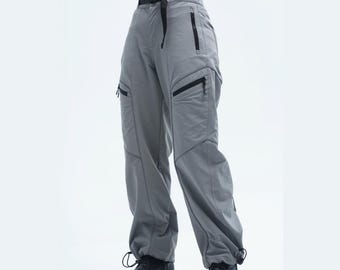 Könnte beinhalten: Graue Cargohose mit mehreren Taschen und schwarzen Reißverschlüssen. Die Hose hat einen schwarzen Gürtel und Kordelzüge an den Knöcheln. Das Model trägt schwarze Stiefel. Die Hose ist aus leichtem Material.