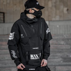 May include: Black anorak jacket with multiple pockets, zippers, and a hood. The jacket features white graphic designs on the sleeves and chest, along with the text "TUR WEIR". The person is wearing a black cap and face mask.