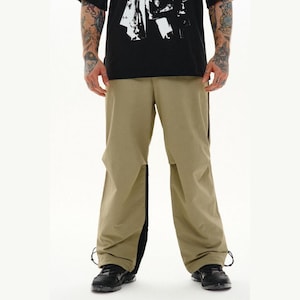 May include: Beige trousers featuring a loose, wide-leg fit with contrasting black side panels running down the length of each leg. The pants include adjustable drawstrings at the hem and are paired with a black t-shirt with a white graphic print.