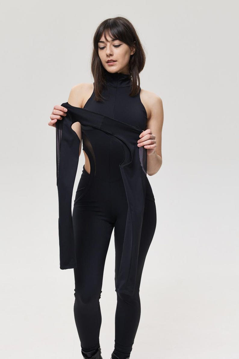 May include: Black jumpsuit with a high neckline and long sleeves. The jumpsuit features cut-out details on the sides and a form-fitting silhouette. The sleeves are attached to the jumpsuit with a mesh panel. The model is holding the sleeve.