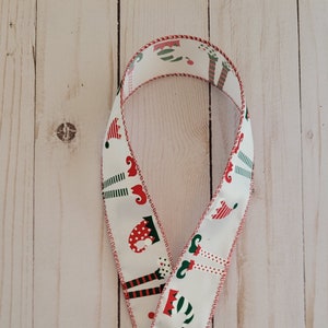 May include: White ribbon with a red stitched edge and a repeating pattern of green, red, and white elf legs and hats. The ribbon is folded in half and the text "Door Decor And Things" is visible in the bottom center of the image.
