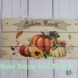 May include: A wooden sign with a distressed white background featuring the text "Autumn Harvest" in a banner with a fall-themed design of pumpkins, sunflowers, and leaves.