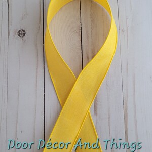 May include: A bright yellow ribbon, folded in half, on a white wood background. The ribbon is a solid color with a slight sheen.
