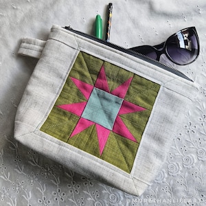 May include: A square, light gray fabric pouch with a starburst design. The starburst is pink and light blue on a green background. The pouch has a black zipper, a small handle, and contains sunglasses and pens.