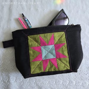 May include: Black zippered pouch with a colorful starburst design. The starburst features pink, green, and light blue geometric shapes. The pouch contains pens and sunglasses. The text "morethanlifeart" is visible.