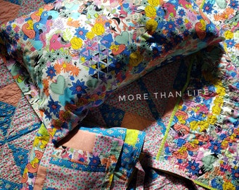 May include: A colorful patchwork quilt with a floral print pillow and a matching tote bag. The pillow has a white background with colorful flowers, hearts, and geometric shapes. The tote bag is made of different patterned fabrics in shades of pink, blue, and green. The quilt has a chevron pattern in shades of pink and blue.