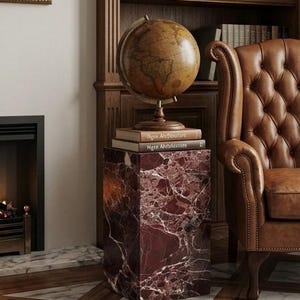May include: A vintage-style globe sits atop a stack of books on a red marble pedestal. The pedestal has white veining. A brown leather armchair and a wooden bookshelf filled with books are in the background. The scene evokes a classic library setting.
