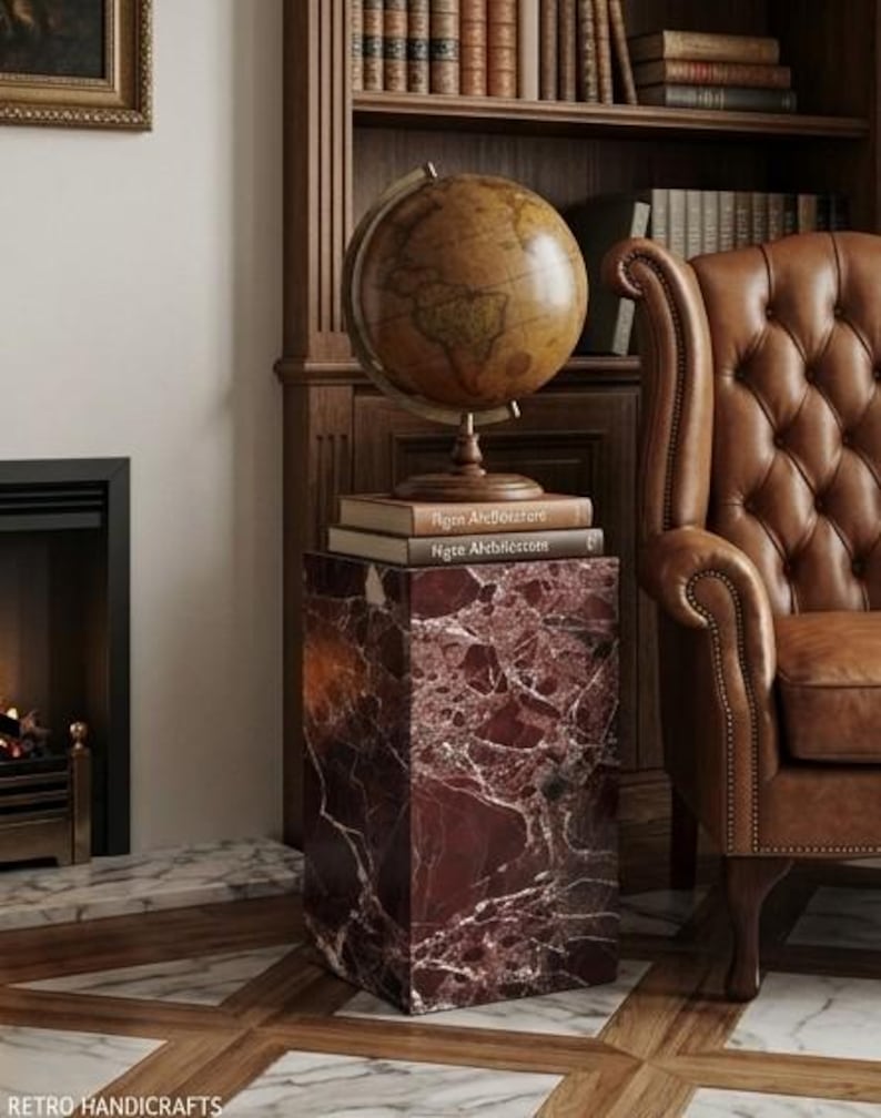 May include: A vintage-style globe sits atop a stack of books on a red marble pedestal. The pedestal has white veining. A brown leather armchair and a wooden bookshelf filled with books are in the background. The scene evokes a classic library setting.