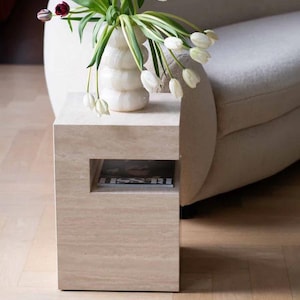May include: A beige stone side table with a rectangular shape and a recessed shelf. A white vase with a ribbed design holds white and purple tulips. The table is next to a curved, off-white sofa.