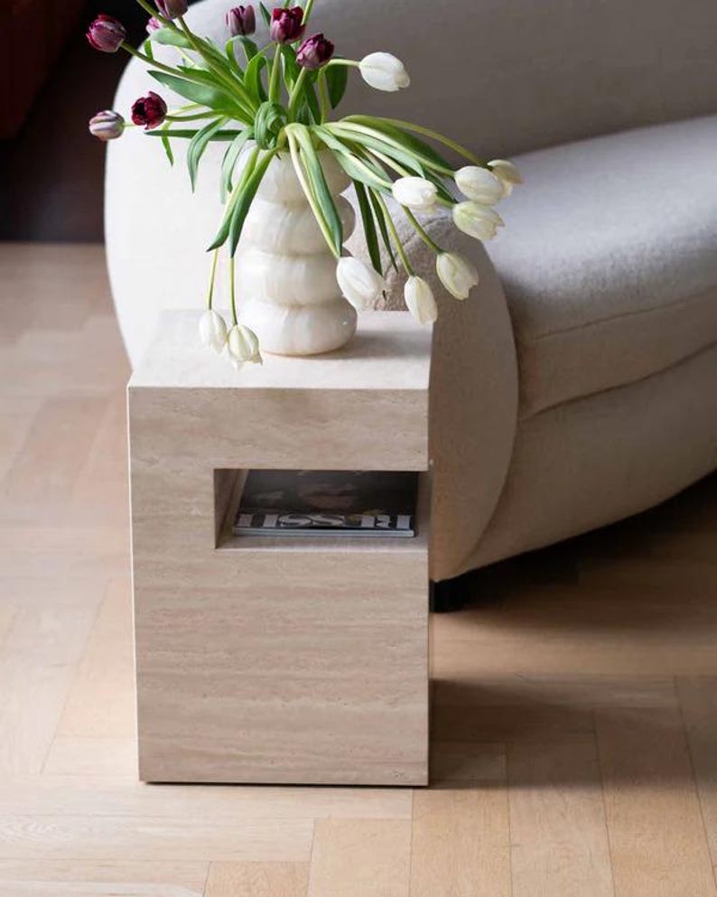 May include: A beige stone side table with a rectangular shape and a recessed shelf. A white vase with a ribbed design holds white and purple tulips. The table is next to a curved, off-white sofa.
