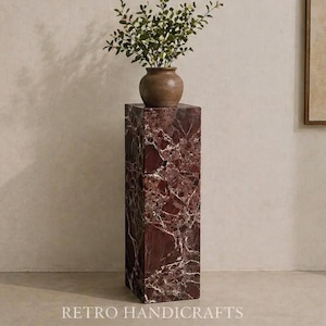 May include: A tall, rectangular pedestal made of red marble with white veining. A small, brown vase with green foliage sits atop the pedestal. The base of the pedestal has the text "RETRO HANDICRAFTS".