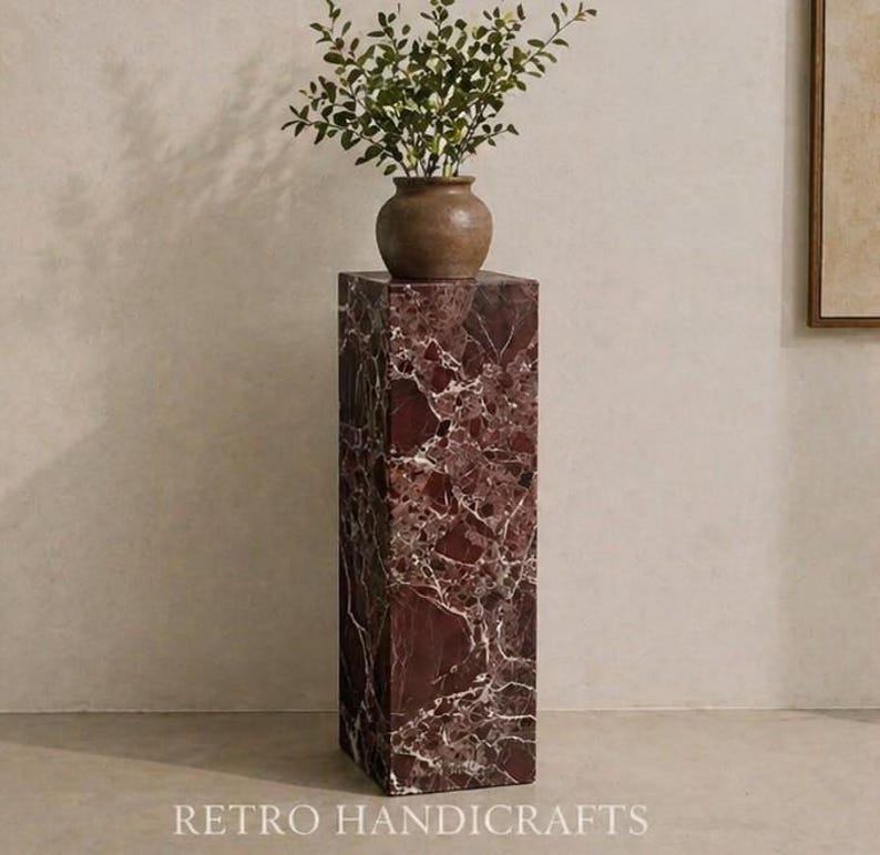 May include: A tall, rectangular pedestal made of red marble with white veining. A small, brown vase with green foliage sits atop the pedestal. The base of the pedestal has the text "RETRO HANDICRAFTS".