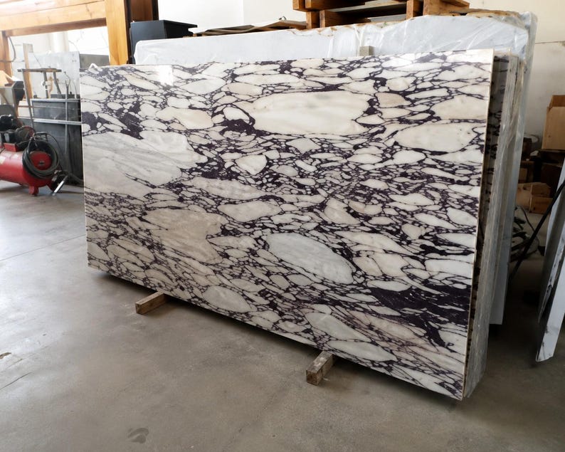 May include: A large rectangular slab of polished stone featuring a high-contrast pattern of irregular white and cream-colored shapes embedded within a dense, dark purple and black veined matrix, standing upright on two small wooden support blocks.