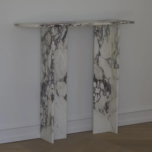 Puede incluir: Una consola de mármol con una parte superior rectangular y dos soportes verticales. El mármol blanco presenta vetas de color gris oscuro. La mesa está colocada contra una pared blanca y se apoya en un suelo de madera.