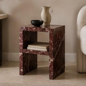 May include: A square side table with a red marble pattern, featuring a lower shelf with books, a black bowl, and a beige vase. The table is next to a cream-coloured armchair.