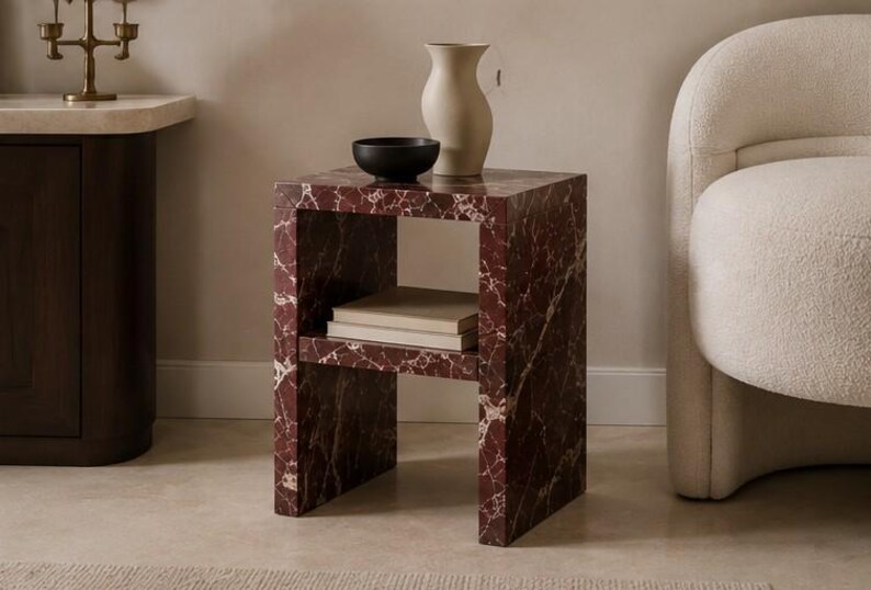 May include: A square side table with a red marble pattern, featuring a lower shelf with books, a black bowl, and a beige vase. The table is next to a cream-coloured armchair.
