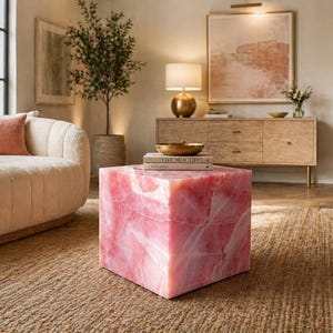 Può includere: Un tavolino quadrato in marmo rosa con venature. Il tavolo si trova in un soggiorno con un divano beige, un mobile in legno e un tappeto intrecciato. Una ciotola dorata e dei libri sono sul tavolo.