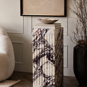 May include: A rectangular marble pedestal featuring vertical fluted grooves and a white base with deep purple veining. Two stacked books titled Live Beautiful and This is Home sit on top, supporting a shallow, wavy-edged beige ceramic bowl.
