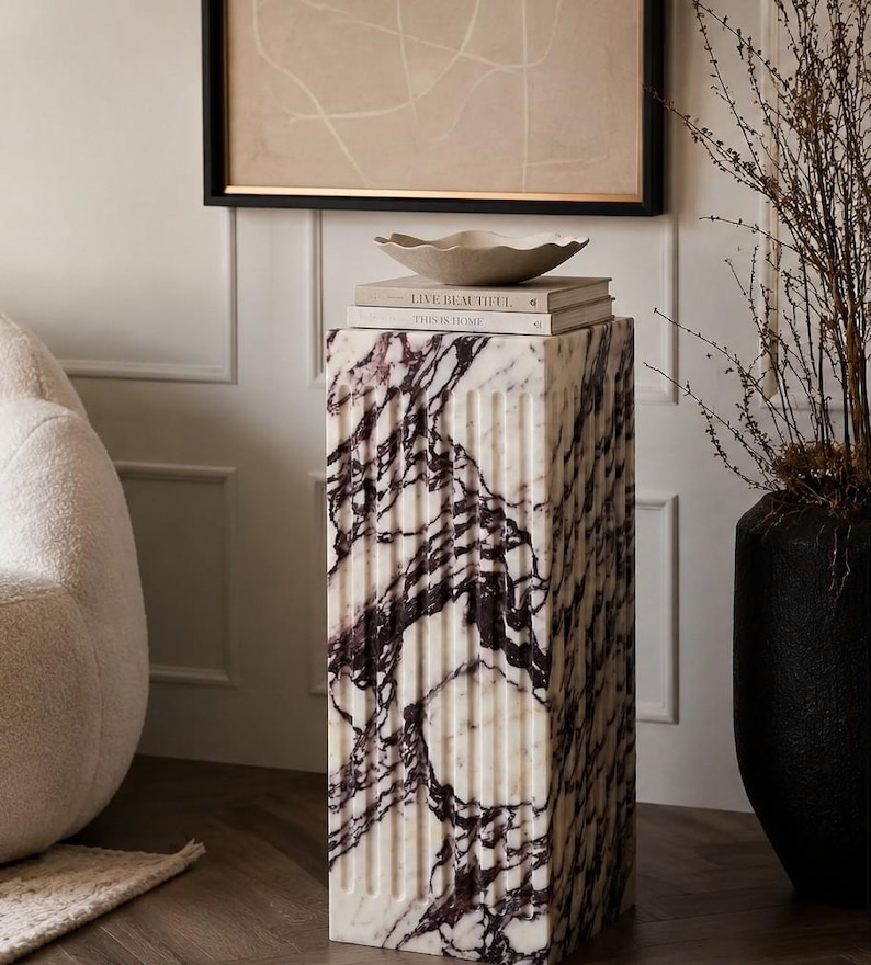 May include: A rectangular marble pedestal featuring vertical fluted grooves and a white base with deep purple veining. Two stacked books titled Live Beautiful and This is Home sit on top, supporting a shallow, wavy-edged beige ceramic bowl.