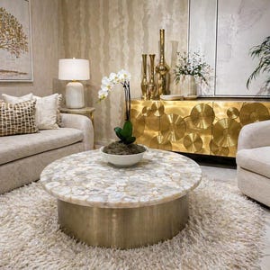 Puede incluir: Un salón con una mesa de centro redonda con una tapa de mosaico y una base dorada. Un sofá beige con cojines está cerca de la mesa. Un mueble dorado y un sillón blanco también están en la habitación.