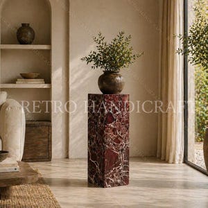 May include: A tall, rectangular, red marble pedestal with white veining supports a dark brown vase filled with green foliage. The pedestal is in a room with a neutral color palette, a sofa, and a large window with a potted tree.