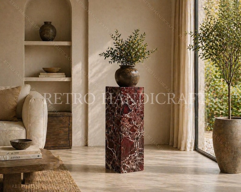 May include: A tall, rectangular, red marble pedestal with white veining supports a dark brown vase filled with green foliage. The pedestal is in a room with a neutral color palette, a sofa, and a large window with a potted tree.