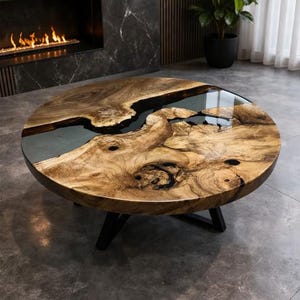 May include: A round coffee table with a distinctive design. The tabletop is made of natural wood with a dark epoxy resin inlay, resembling a river. The table has a dark, three-legged base. The wood grain is visible, and the table has a glossy finish.