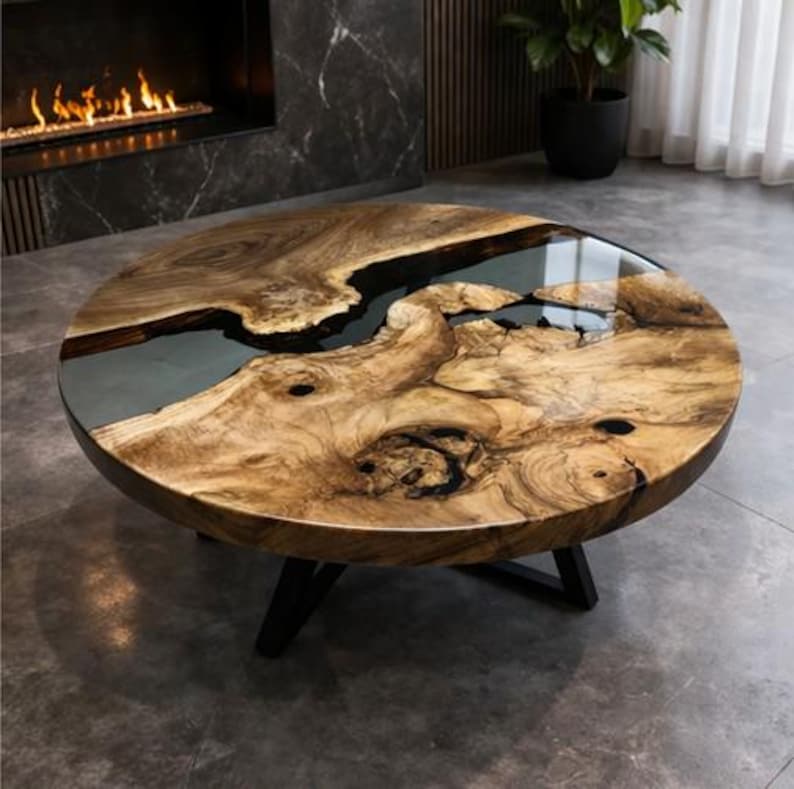 May include: A round coffee table with a distinctive design. The tabletop is made of natural wood with a dark epoxy resin inlay, resembling a river. The table has a dark, three-legged base. The wood grain is visible, and the table has a glossy finish.