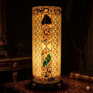 Puede incluir: Una lámpara cilíndrica con una pantalla de color crema con intrincados patrones calados y motivos florales y animales coloridos. La lámpara tiene una base oscura y está iluminada desde adentro, creando un brillo cálido. El diseño incluye un mono, un pavo real y un pájaro.