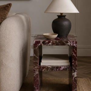 May include: A side table with a deep red marble finish and a lower shelf. A black ceramic lamp with a beige lampshade sits on top, along with a small beige bowl. The table is next to a beige sofa.