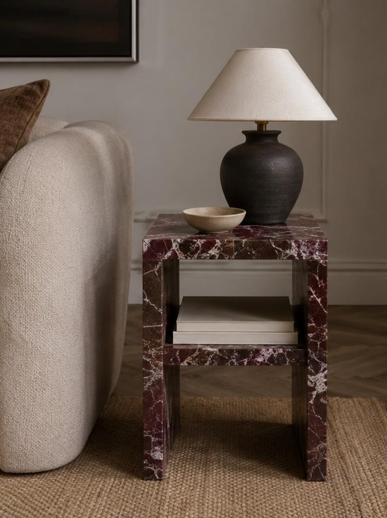 May include: A side table with a deep red marble finish and a lower shelf. A black ceramic lamp with a beige lampshade sits on top, along with a small beige bowl. The table is next to a beige sofa.