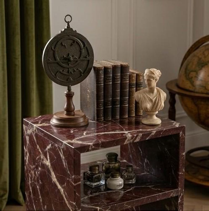 May include: A red marble side table displaying antique books, a bust, and a vintage astrolabe. The table also features small glass bottles. The table's dimensions are not visible, but the items suggest a classic, historical theme.