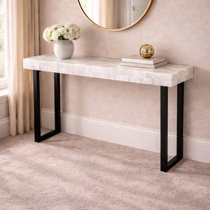 May include: A white console table with a mosaic-like top and black metal legs. A round gold mirror hangs on the wall above the table. A white vase with white flowers and decorative items are on the table.