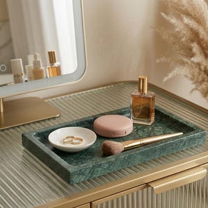 Può includere: Un tavolo da toeletta con un vassoio in marmo verde contenente un flacone di profumo, un pennello per il trucco, un compatto e un piattino bianco con anelli d'oro. Uno specchio con cornice dorata ed erba della pampa decorativa sono sullo sfondo.
