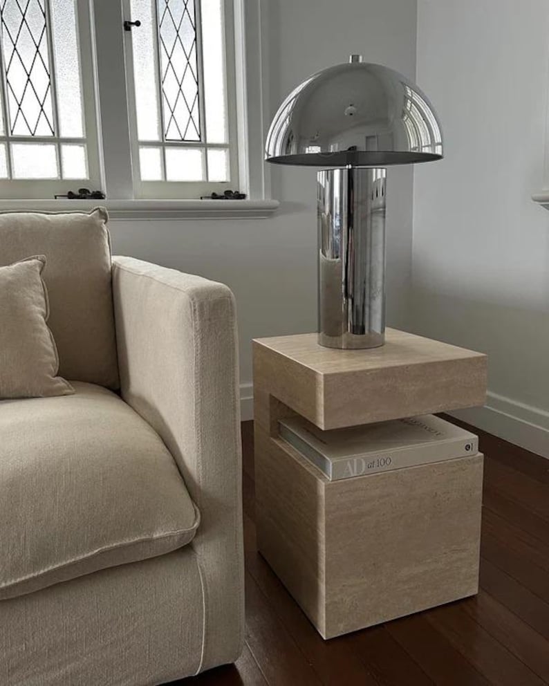 May include: A modern chrome mushroom-shaped table lamp sits atop a light-colored stone side table. The lamp has a cylindrical base and a rounded shade. The side table has a unique design with a recessed shelf holding a book. A beige sofa is in the background.